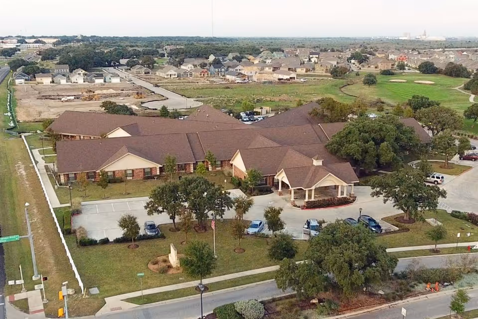 Aerial view of Legend Oaks Rehabilitation and Healthcare - Kyle, showing a large single-story building with a brown roof surrounded by trees, parking lots with several cars, and green lawns. The facility is located in a suburban area with residential houses and open fields in the background.