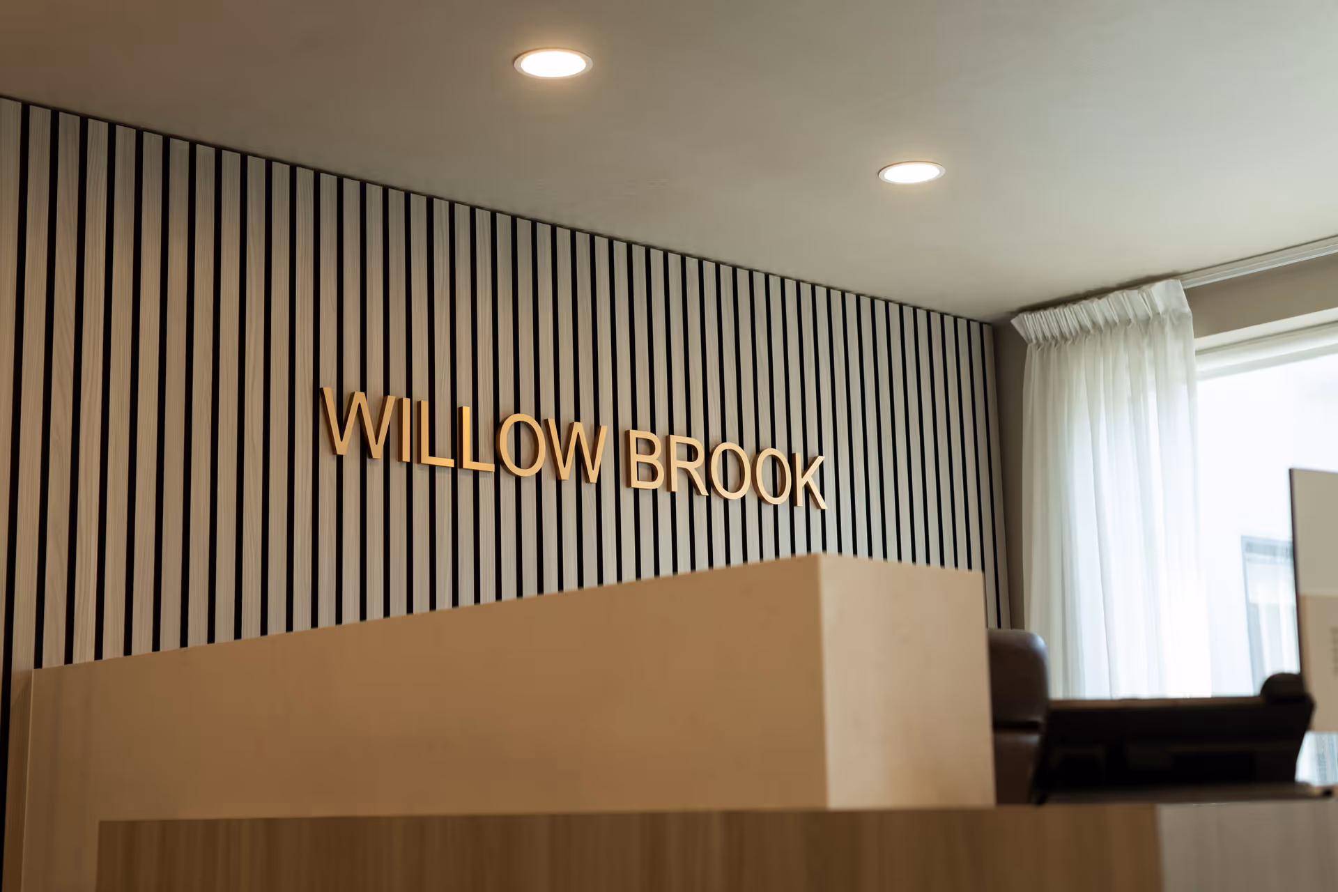 Reception area with a modern wooden slat wall featuring the text 'WILLOW BROOK' in gold letters, a beige reception desk in the foreground, and a window with white curtains allowing natural light into the space.