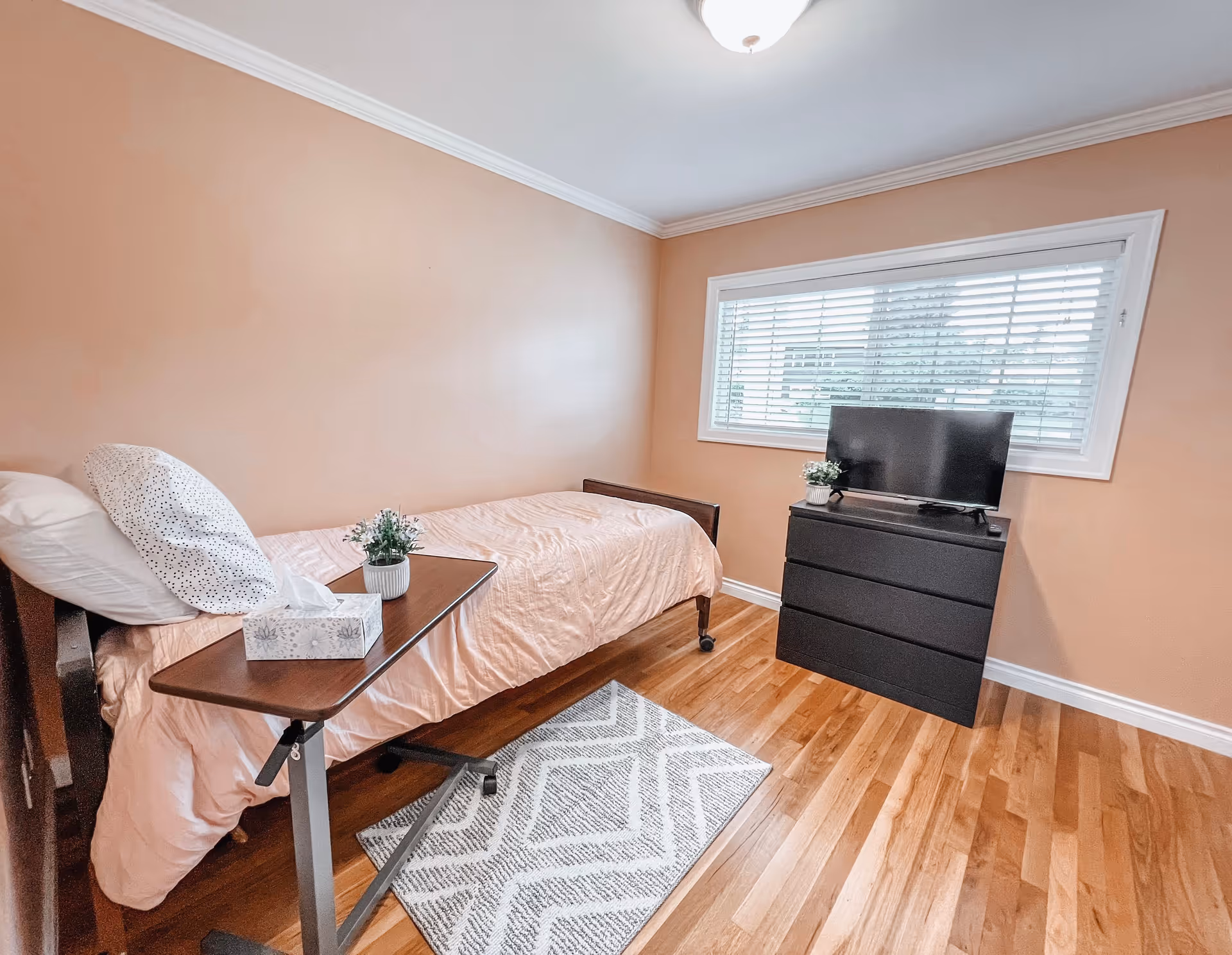 A small bedroom with light peach walls and wooden flooring. The room features a single bed with light pink bedding and two pillows, a small overbed table with a tissue box and a small potted plant, a black dresser with a flat-screen TV on top, and a window with white blinds letting in natural light. A gray and white patterned rug is placed on the floor beside the bed.