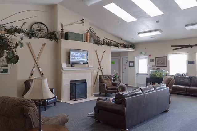 A cozy living room area in Ashley Manor Memory Care featuring leather sofas and armchairs arranged around a fireplace with a mounted TV above it. The room has a high ceiling with skylights, decorative vintage skis and greenery on the walls, and a person sitting on one of the sofas watching TV.
