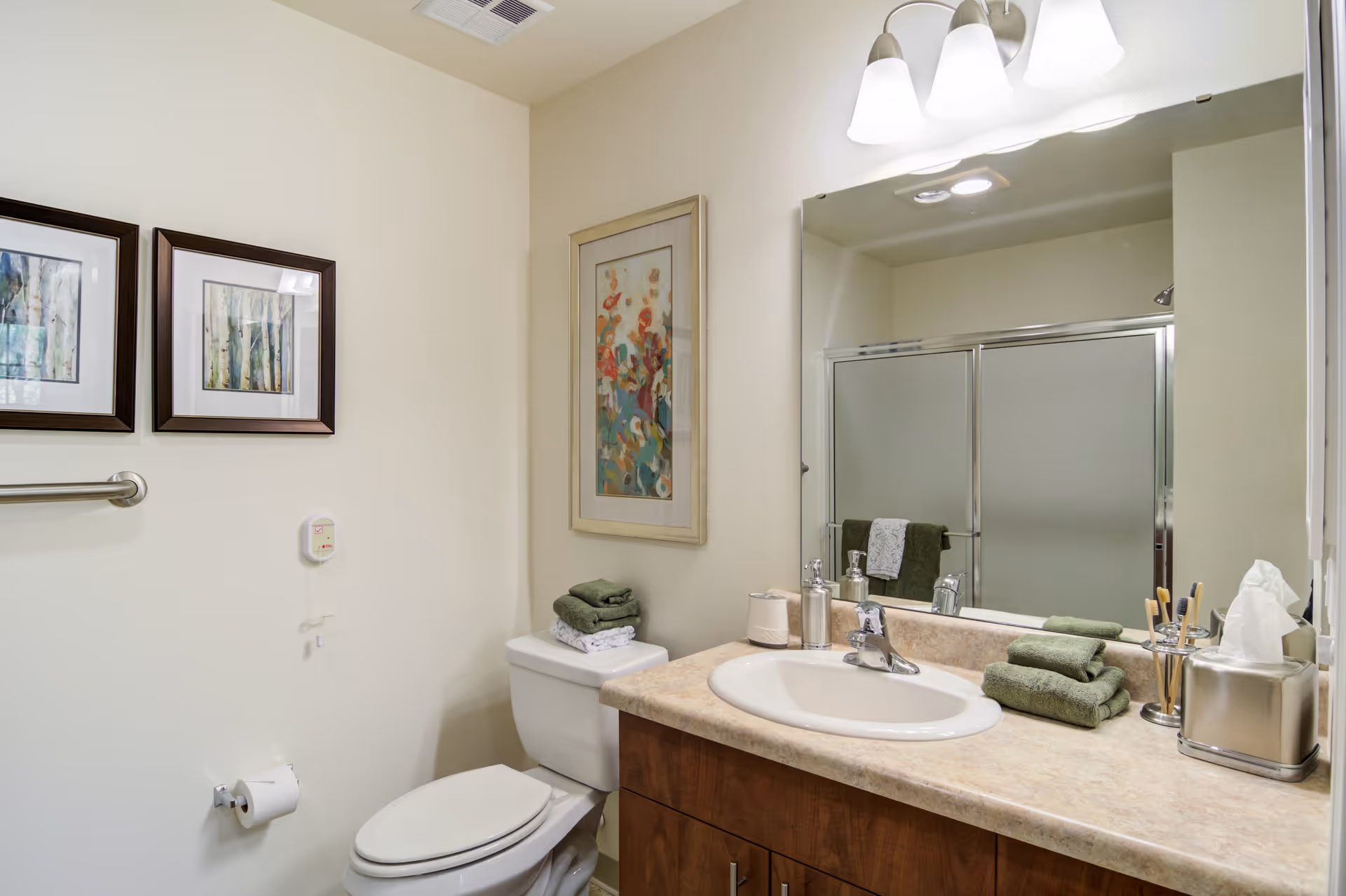 A clean and well-lit bathroom featuring a toilet with folded green towels on top, a countertop with a sink, soap dispenser, tissue box, and toothbrush holder. Above the sink is a large mirror with three light fixtures. The shower has frosted glass sliding doors. The walls are decorated with framed artwork.