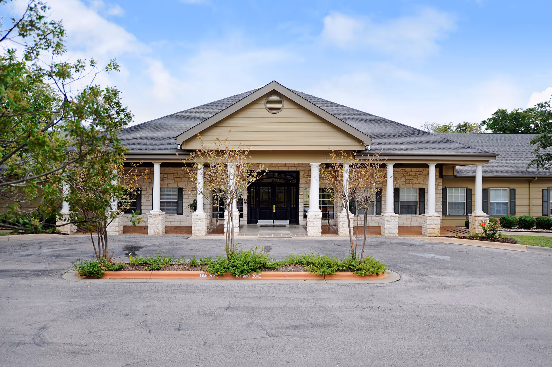 Front entrance of a single-story senior living facility with a covered portico supported by columns, stone facade, and a driveway with landscaping.