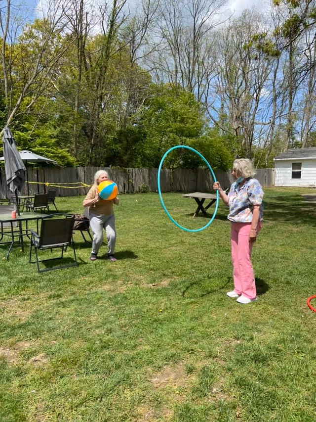 Two older women play with a beach ball and a large hula hoop in a grassy fenced yard with patio furniture and trees.