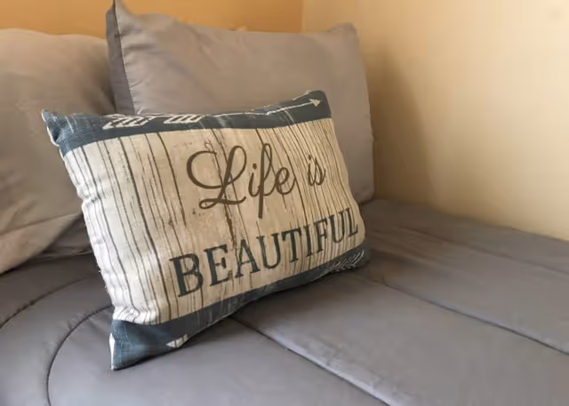 A close-up view of a bed with gray bedding and pillows, featuring a decorative pillow with the text 'Life is BEAUTIFUL' on it.