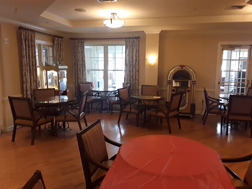 A cozy dining area with several round tables and chairs arranged on a wooden floor. One table in the foreground is covered with a red tablecloth. The room has large windows with patterned curtains, a popcorn machine, and a vintage jukebox against the wall. Warm lighting fixtures illuminate the space.
