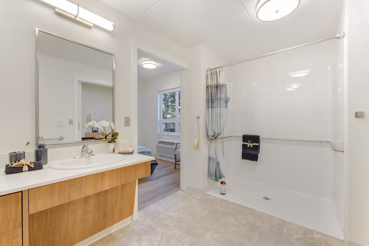 A bright and clean bathroom with a large mirror above a wooden vanity with a white sink. To the right, there is a walk-in shower with a grab bar and a shower curtain partially pulled to one side. A small towel hangs on the grab bar. The bathroom opens into a bedroom with a window and an air conditioning unit visible.