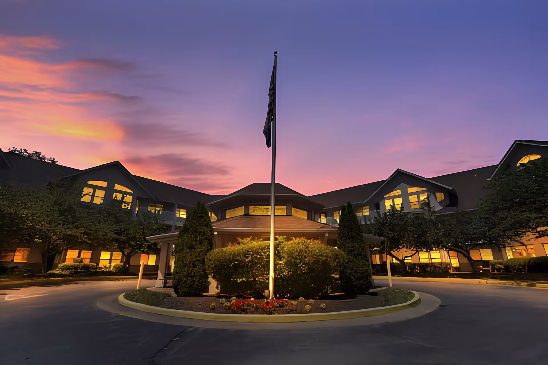 Front entrance of a large senior living building at dusk with a flagpole and illuminated windows.