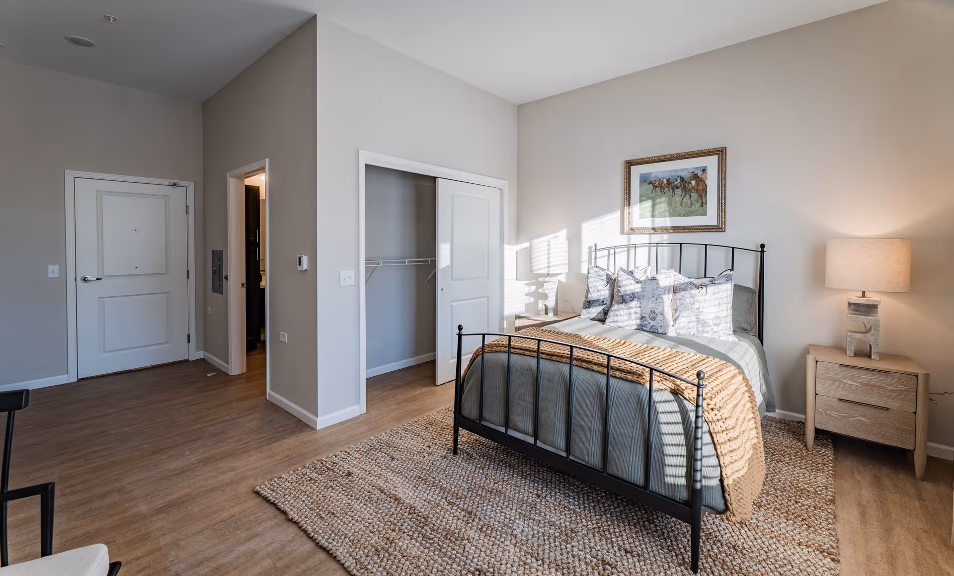 A bright and clean bedroom with a metal framed bed dressed in gray and beige bedding, a textured area rug, a wooden nightstand with a lamp, a framed picture of horses on the wall, an open closet with sliding doors, and a door leading to another room.