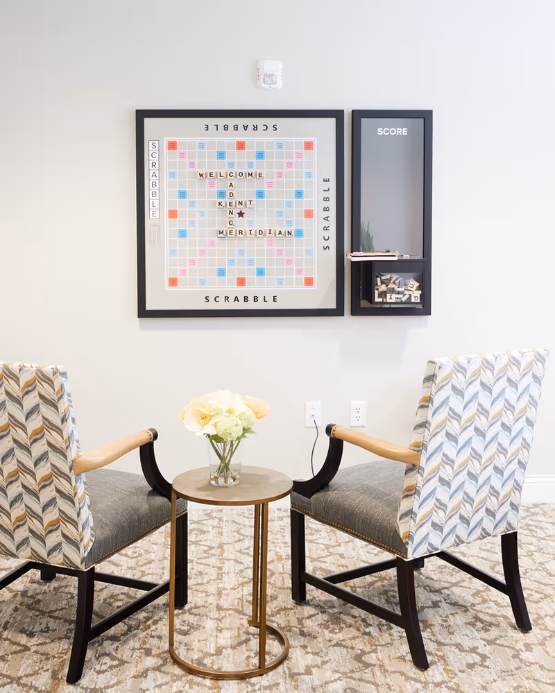 Two upholstered chairs flank a small round table with a vase of flowers beneath a framed Scrabble board and score board on a light wall.