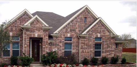Front exterior view of a single-story brick house with multiple gables, several windows, a front door with a small porch, and landscaped bushes and flowers in front.