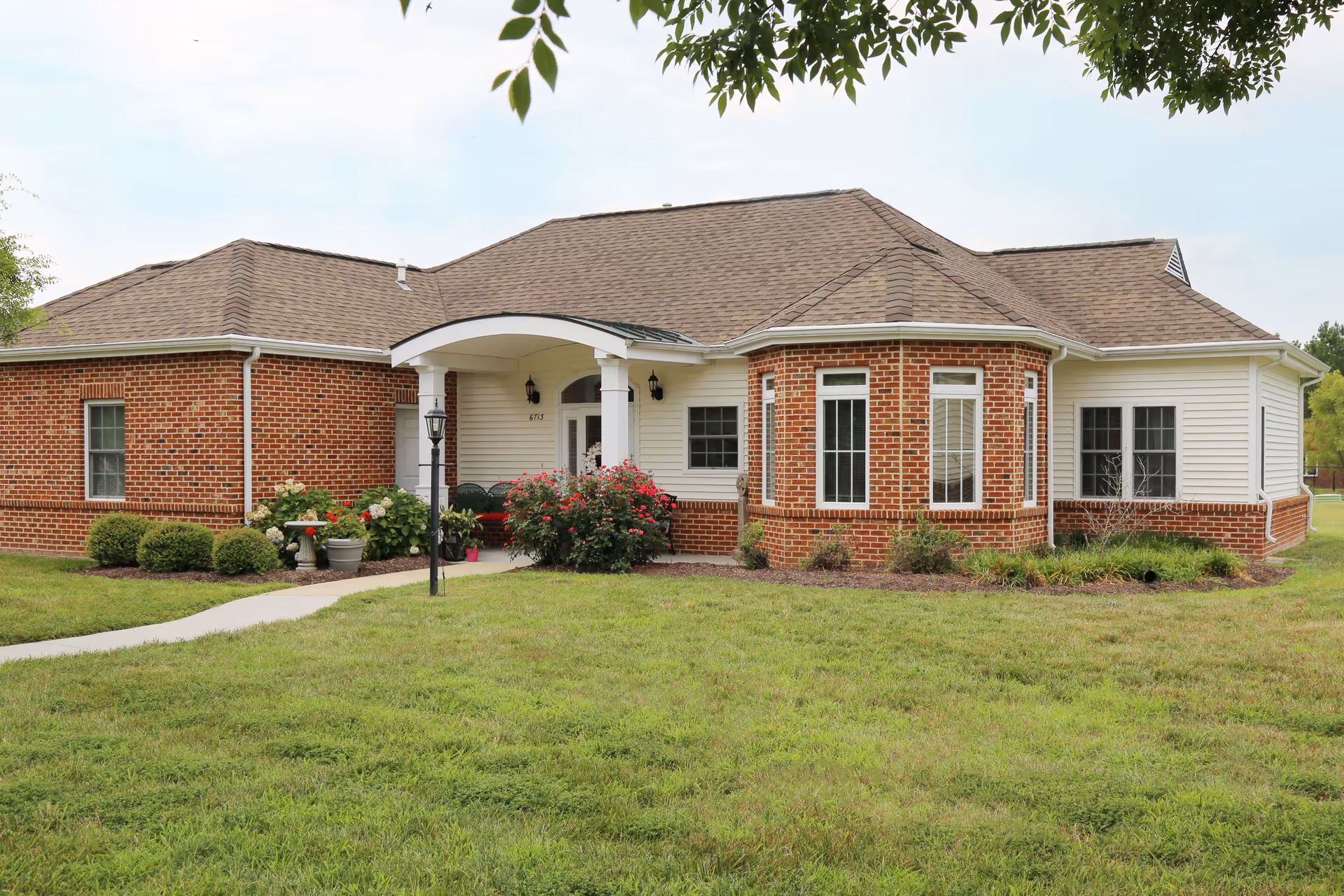 Exterior view of a single-story brick and siding building with a covered entrance, surrounded by green grass and landscaping with bushes and flowers.