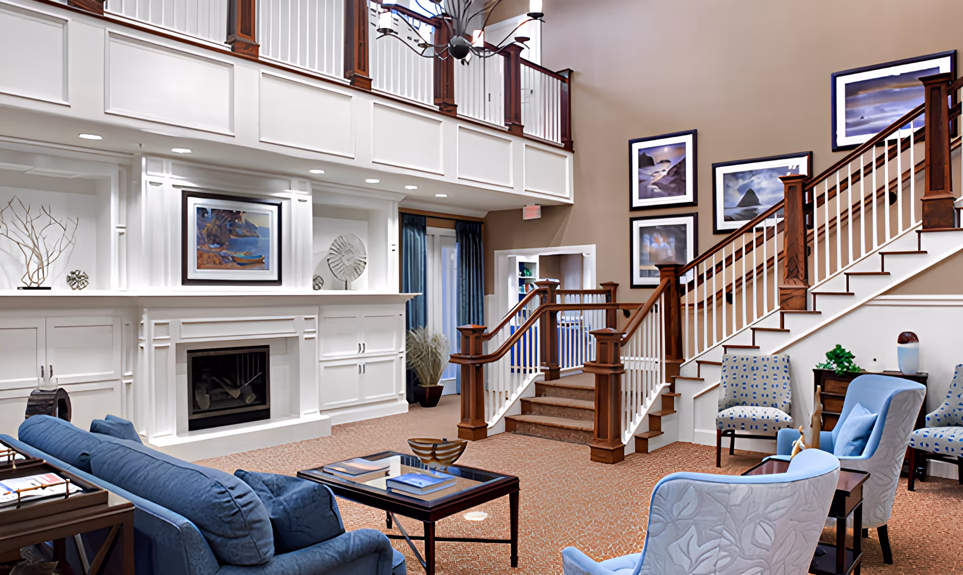 A spacious and elegant living room area in a retirement community featuring a blue sofa, patterned armchairs, a coffee table with books, a fireplace with white built-in cabinetry, and a staircase with wooden railings. The walls are decorated with framed landscape paintings, and there is a balcony railing above the fireplace area.
