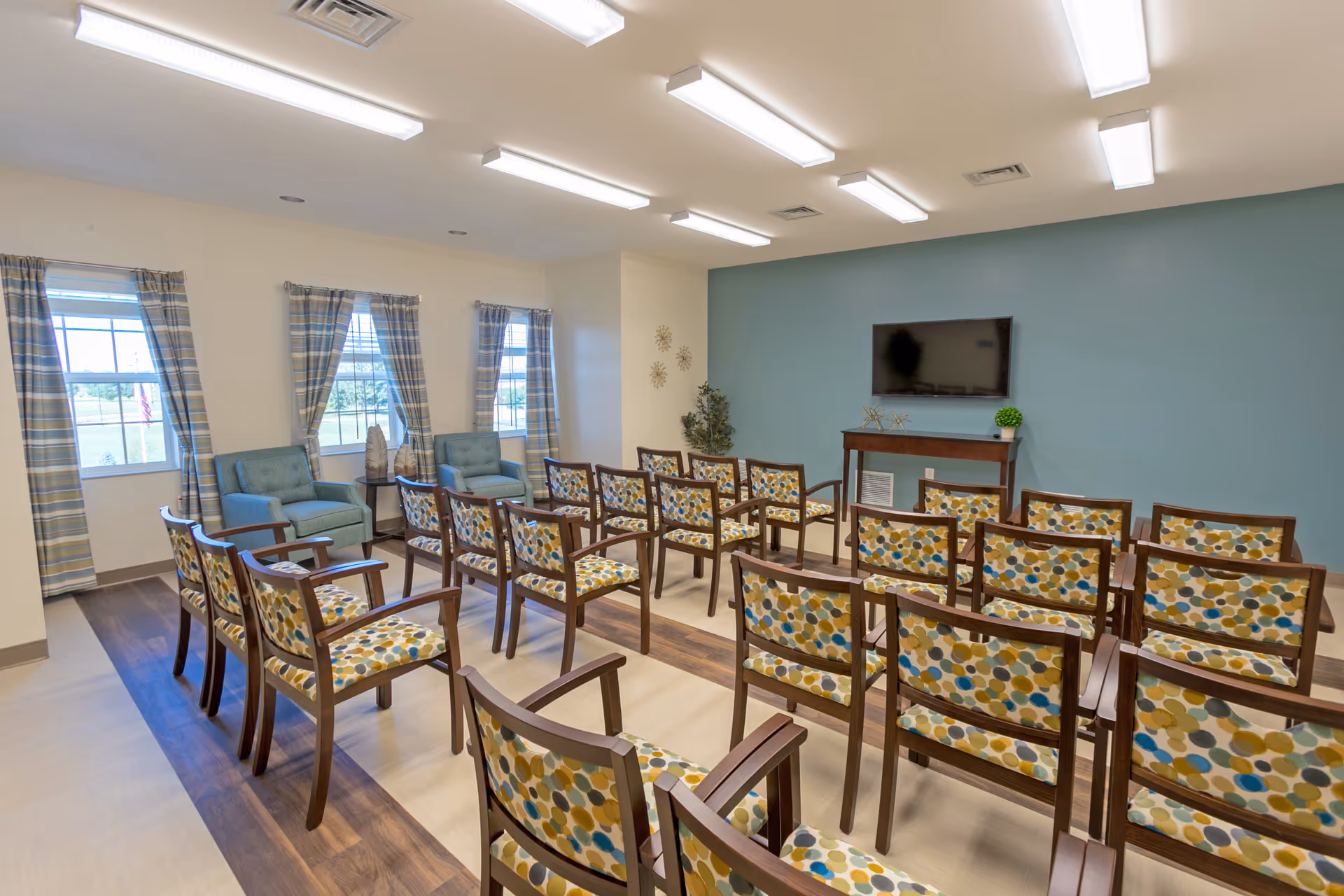 A well-lit meeting or activity room with rows of wooden chairs featuring colorful patterned cushions facing a wall-mounted flat screen TV. The room has three windows with plaid curtains, two blue armchairs near the windows, and a small table with decorative items. The walls are painted white and teal, and the floor has a wood and light-colored pattern.