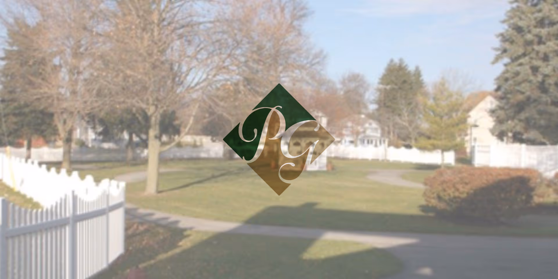 A landscaped outdoor area with a paved walking path, green grass, trees without leaves, bushes, and white fencing surrounding the space. Residential houses are visible in the background under a clear sky.