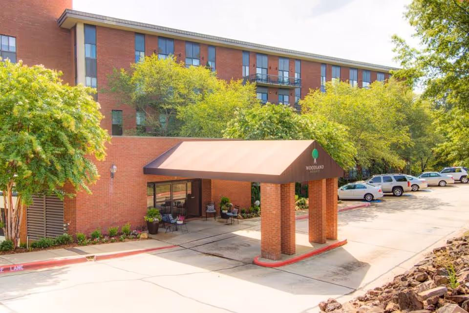 Exterior view of Woodland Heights Senior Living facility showing a brick building with multiple windows, a covered entrance with the facility's name and logo, surrounded by trees and a parking area with several cars.