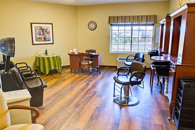 Interior view of a salon area in a senior living facility with several black salon chairs, a wooden desk with a chair, a small table covered with a green cloth, a wall clock, a framed floral picture, and a large window with striped valance letting in natural light.