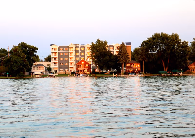 View of a multi-story residential building and several smaller houses along a lakeshore with trees, seen from across the water during sunset.