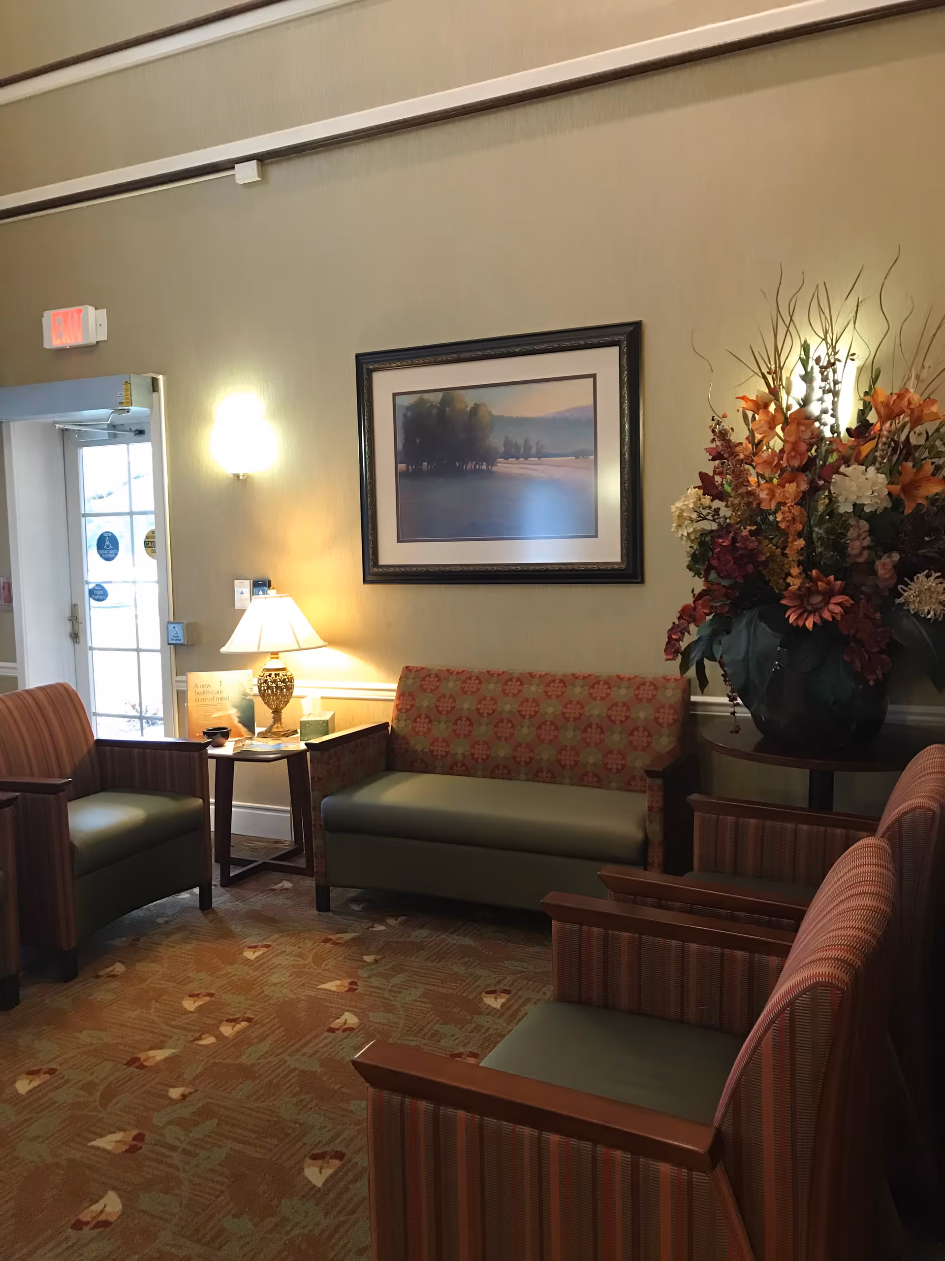 A cozy seating area in a senior living facility with a patterned carpet and four upholstered chairs arranged around a small wooden table with a lamp. A framed landscape painting hangs on the wall above a loveseat, and a large floral arrangement is placed on a side table. A glass door with an exit sign is visible in the background.
