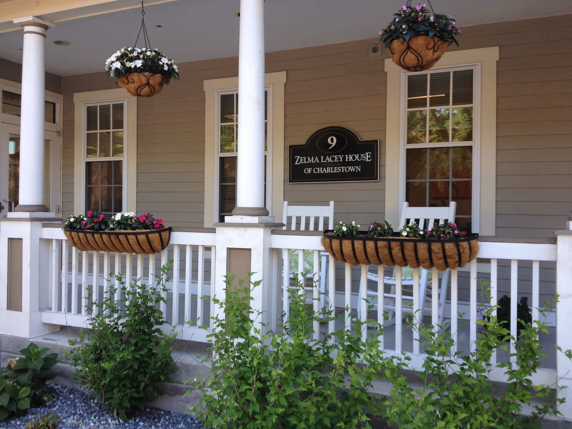 Front porch of a building with beige siding and white trim, featuring two white rocking chairs, hanging flower baskets with white and pink flowers, and a sign that reads '9 Zelma Lacey House of Charlestown'. There are green bushes and plants in front of the porch.