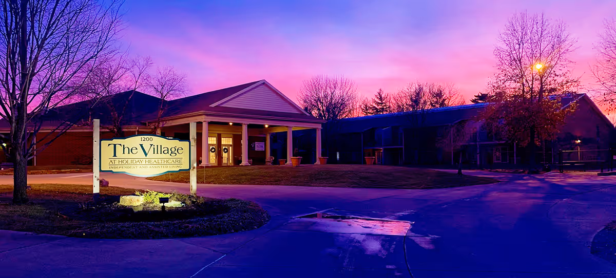 Exterior view of The Village at Holiday Healthcare building during twilight with a purple and pink sky. The building has a covered entrance with columns and a sign in front that reads 'The Village at Holiday Healthcare Independent and Assisted Living'. Leafless trees and a lit streetlamp are visible around the building.