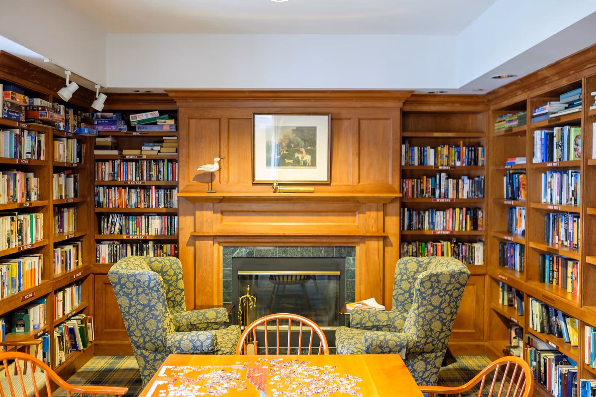 Cozy library room with wooden bookshelves filled with books on three walls, two patterned armchairs facing a wooden fireplace with a framed picture above it, and a wooden table with chairs in the foreground with a partially completed puzzle on it.
