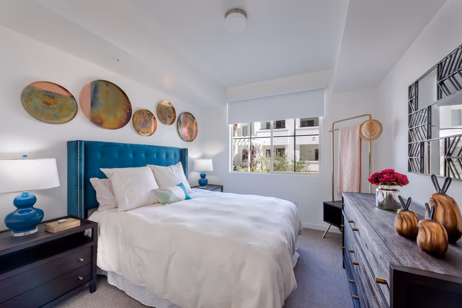 A bright bedroom with a large bed featuring a teal upholstered headboard and white bedding. There are two bedside tables with matching blue lamps. Above the bed, there are five decorative round wall art pieces. A window with a white roller shade lets in natural light. On the right side, there is a dark wooden dresser with decorative items and a mirror above it, along with a gold clothing rack holding a pink towel and a hat.