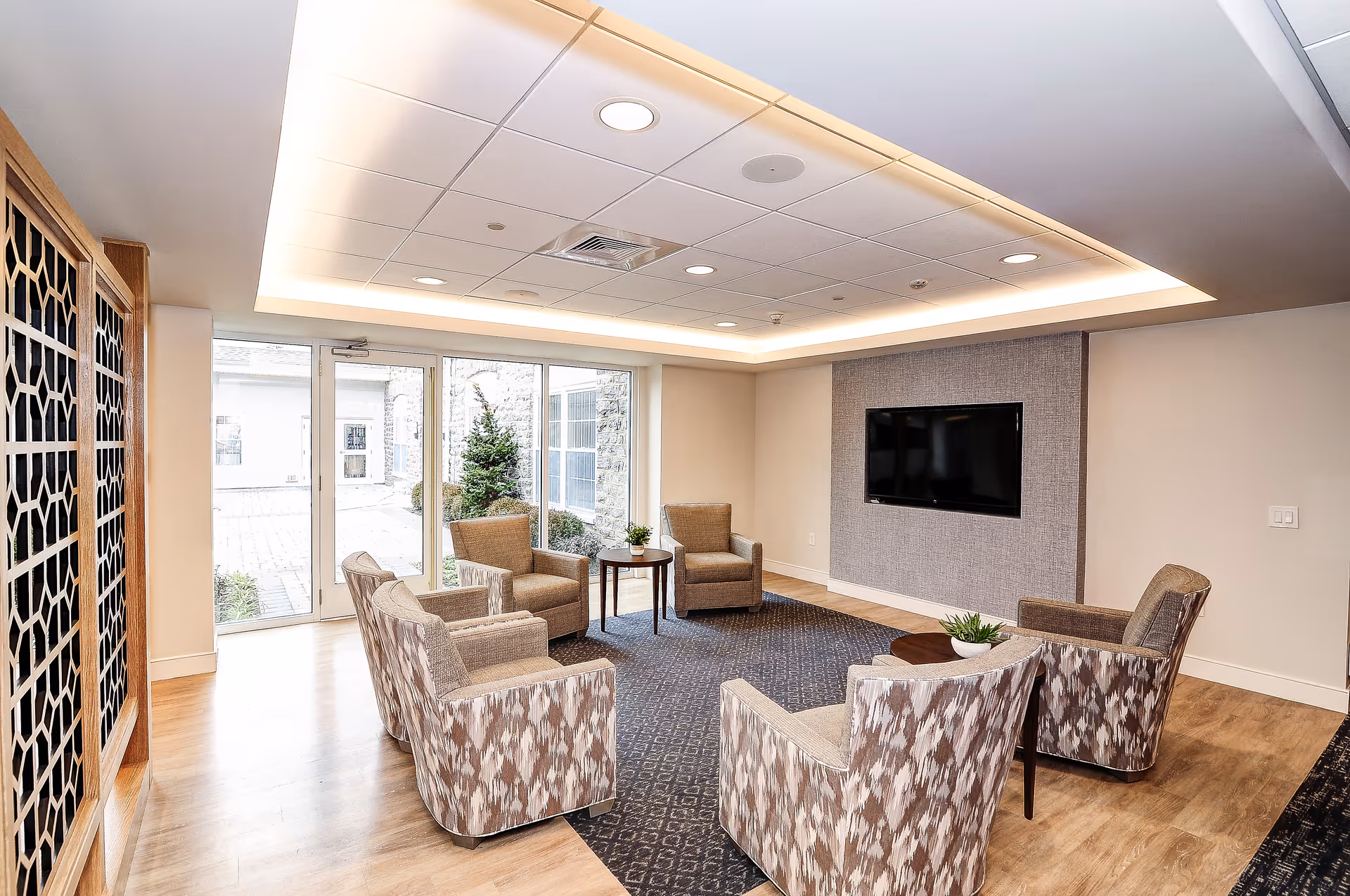 A modern lounge area with six patterned armchairs arranged in a circle around two small round tables with plants. The room has a large window and glass door letting in natural light, a mounted flat-screen TV on a textured wall, light wood flooring, and a ceiling with recessed lighting.