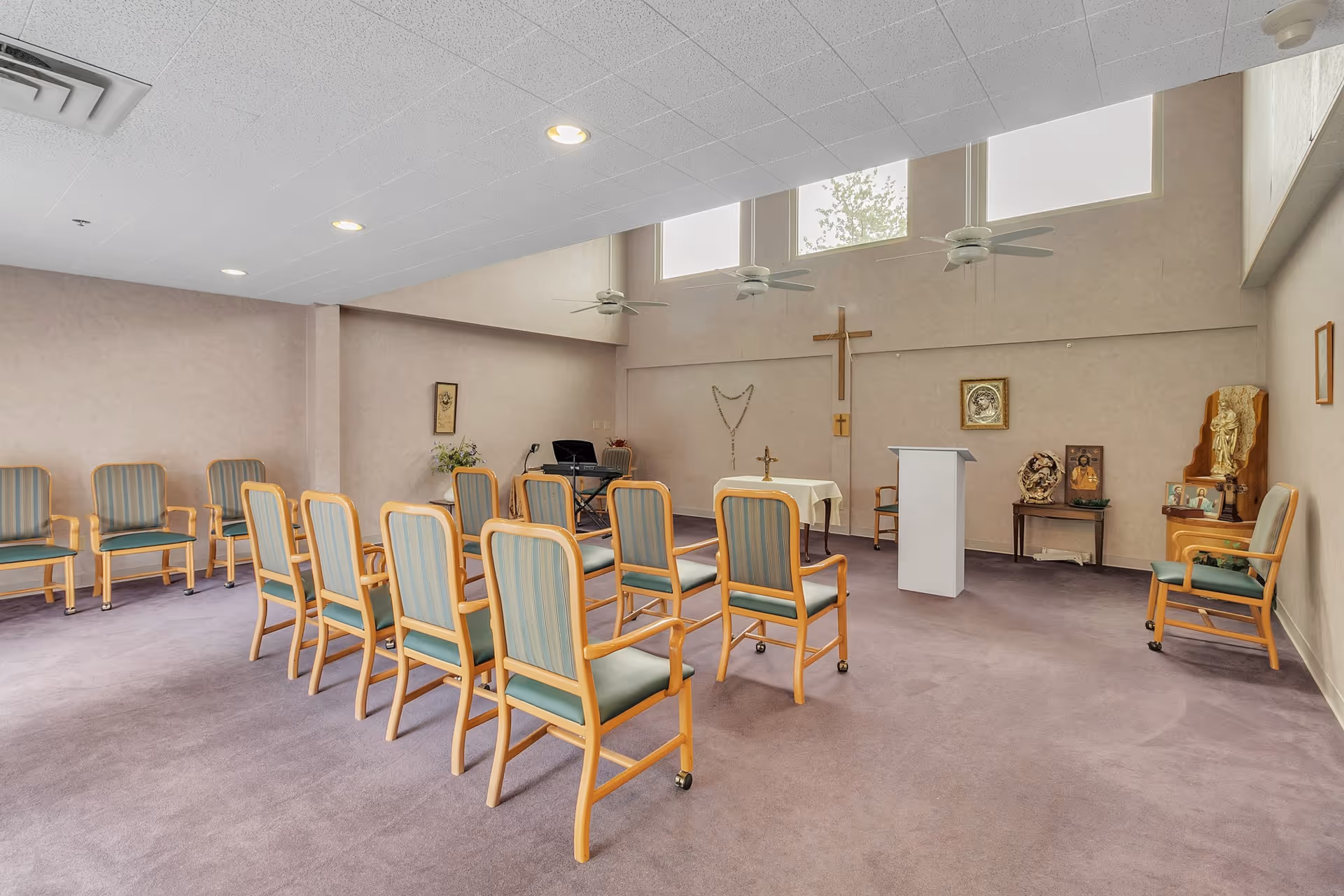 A small chapel or prayer room with rows of wooden chairs with green cushions arranged facing a small altar table with a cross and a lectern. The room has high windows near the ceiling, ceiling fans, and religious decorations including a large wooden cross on the wall, framed pictures, and statues.