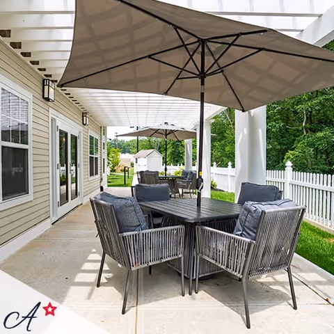 Outdoor patio area with a black metal table and six cushioned chairs under a large beige umbrella. The patio is adjacent to a beige building with white-framed windows and doors, and a white pergola overhead. In the background, there is a white picket fence and green trees.