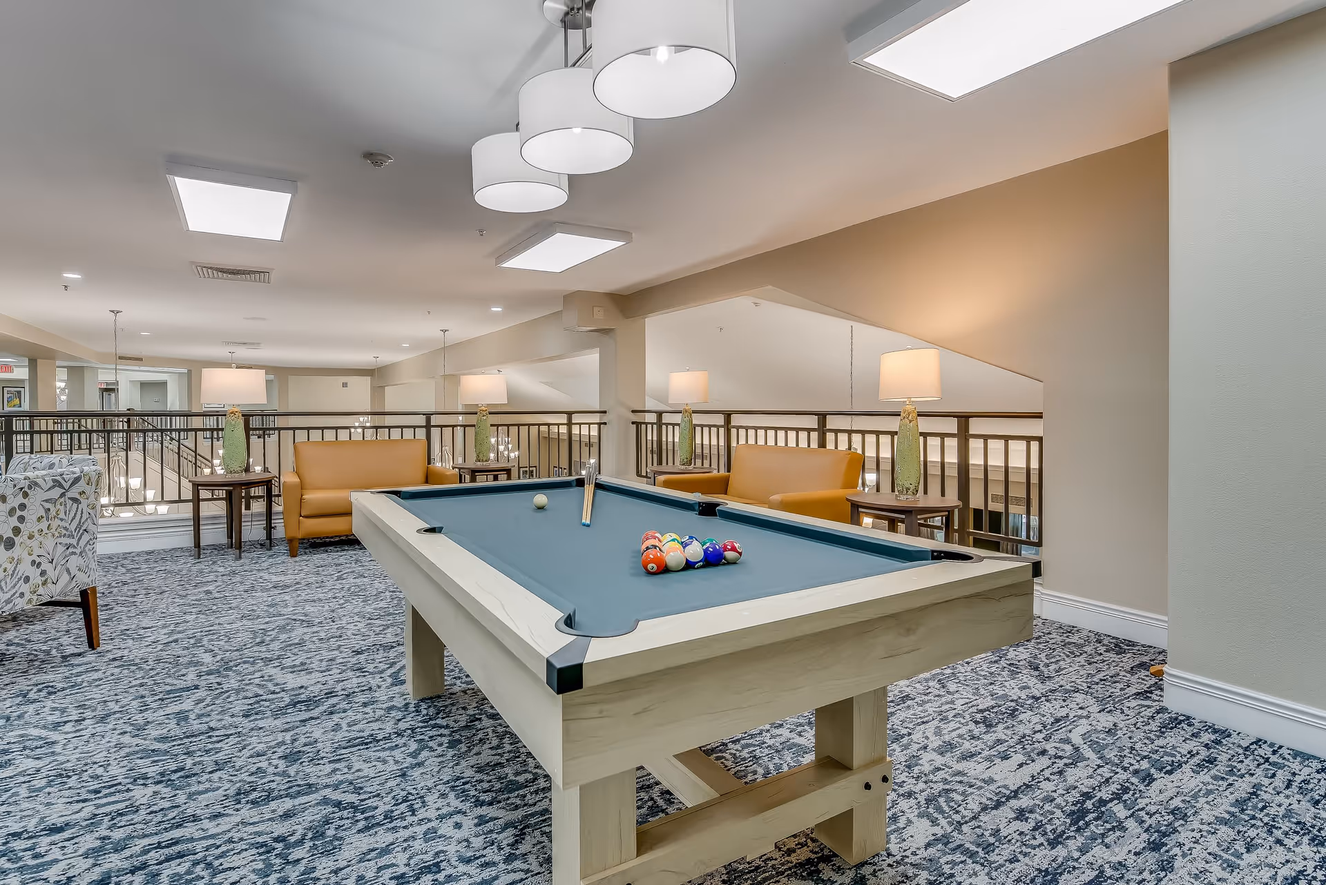 A well-lit recreational room featuring a pool table with billiard balls arranged on it. The room has patterned carpet flooring, beige walls, and modern ceiling lights. There are two mustard-colored sofas with side tables and lamps behind the pool table, and a floral-patterned armchair is partially visible on the left side.