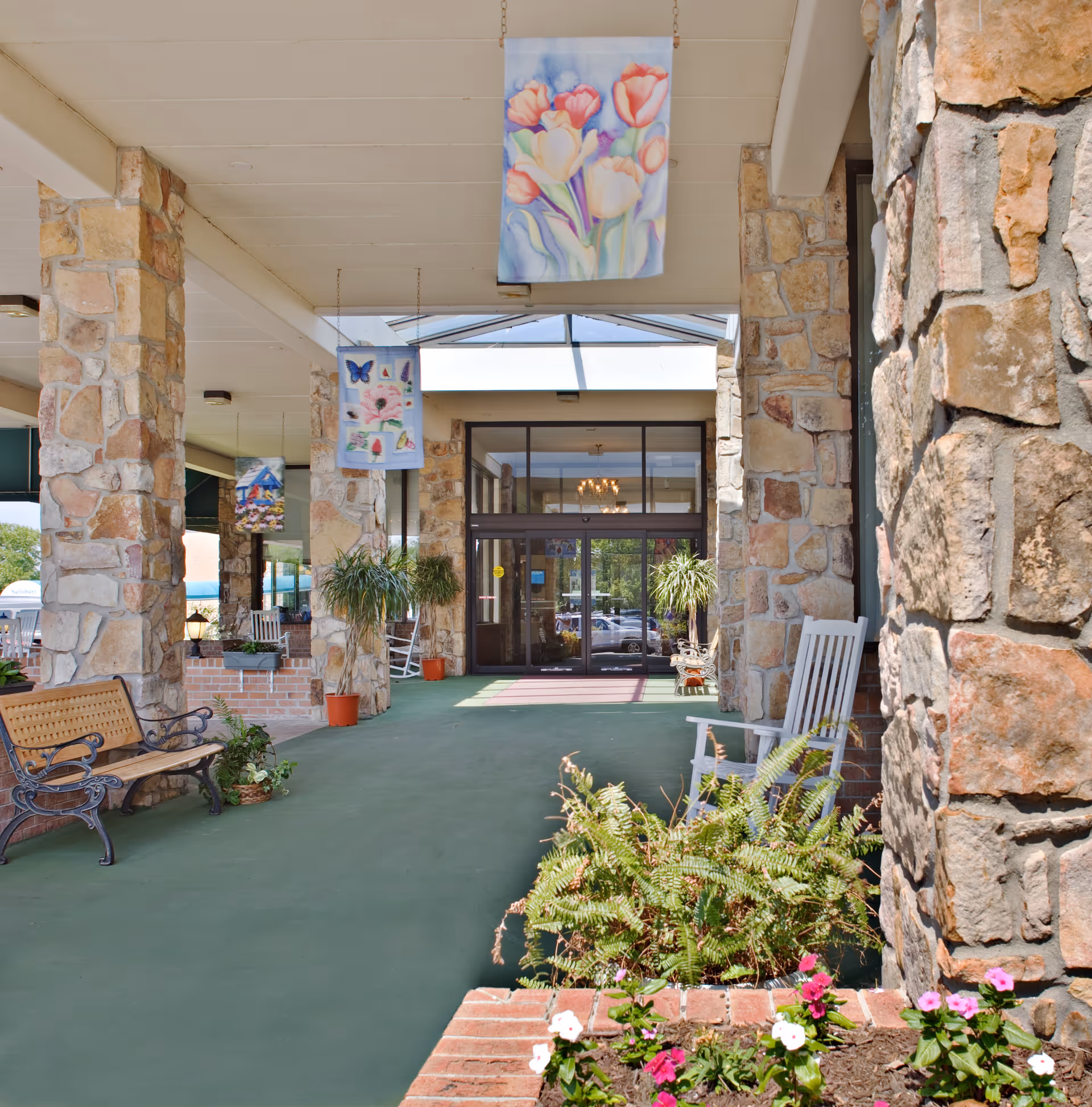 Covered entrance area of a building with stone pillars, hanging decorative banners featuring flowers and butterflies, benches, potted plants, and a glass door entrance at the far end.