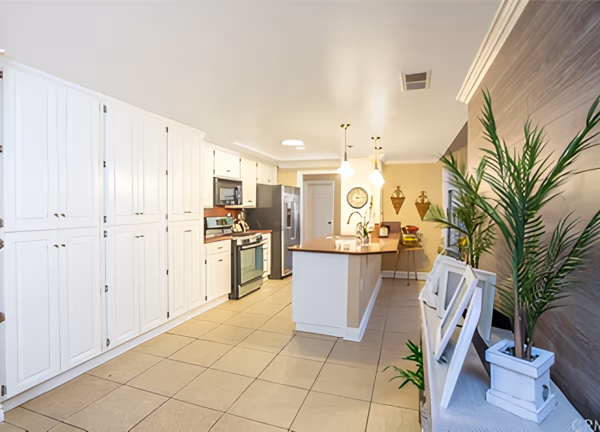 Bright and spacious kitchen with white cabinetry, stainless steel appliances including a refrigerator and stove, a large island with a wooden countertop, pendant lights hanging above the island, and decorative plants and picture frames on a side table.