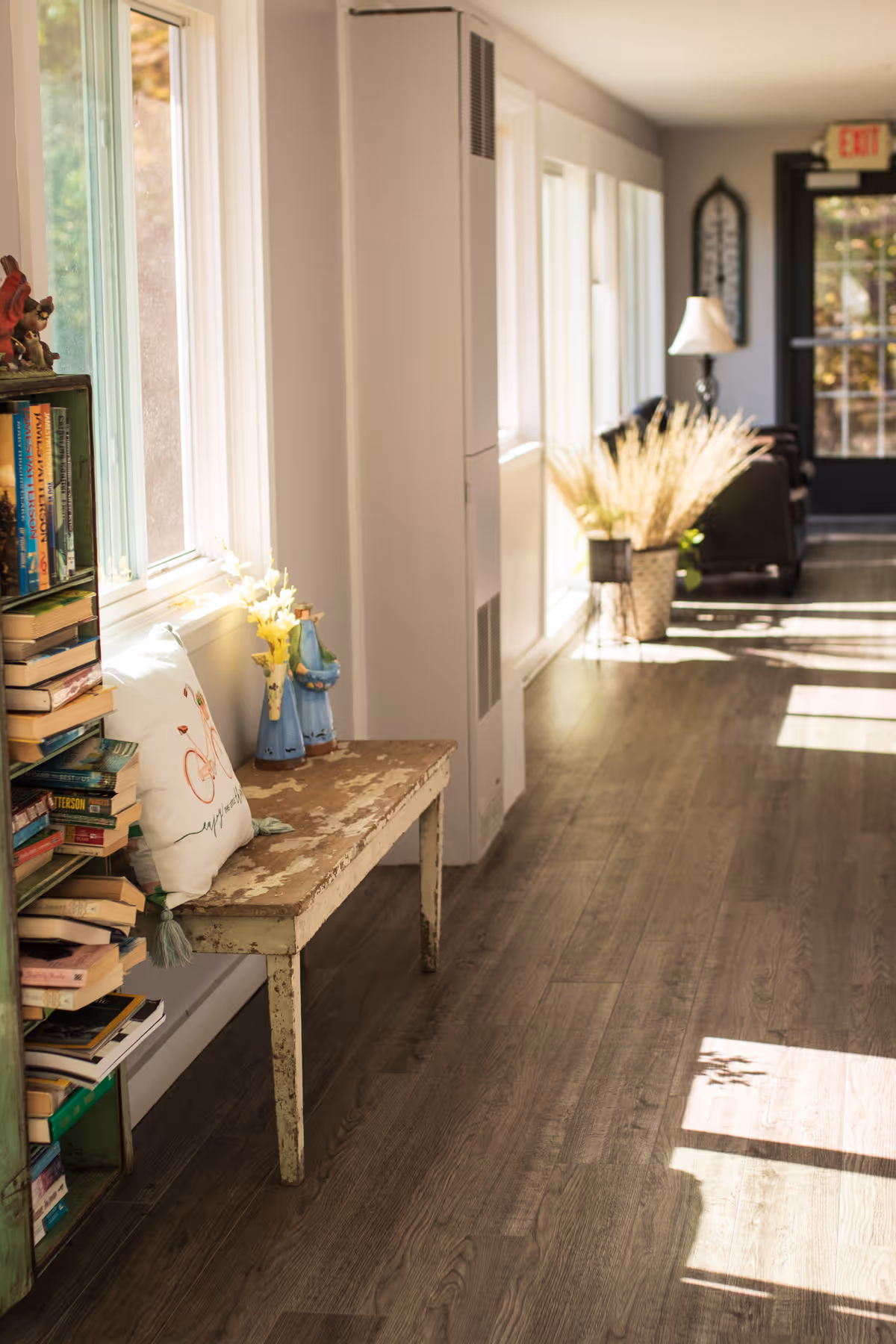 A sunlit hallway in a senior living facility with wooden flooring, a rustic bench with decorative items and a pillow, a bookshelf filled with books, and a seating area with a lamp and potted plants near a glass door.