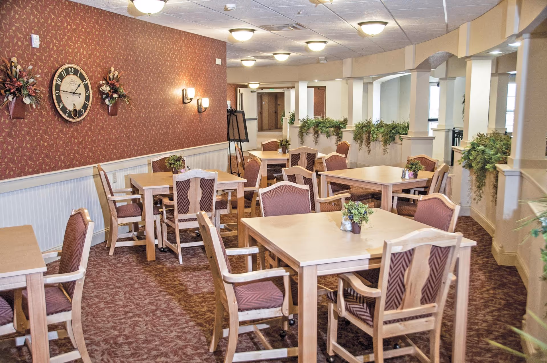 A dining room in a retirement community with several wooden tables and cushioned chairs arranged neatly. The room features a patterned carpet, red wallpaper with floral designs, wall-mounted light fixtures, a large clock, and decorative plants on the walls and tables. The space is well-lit with ceiling lights and has a warm, inviting atmosphere.