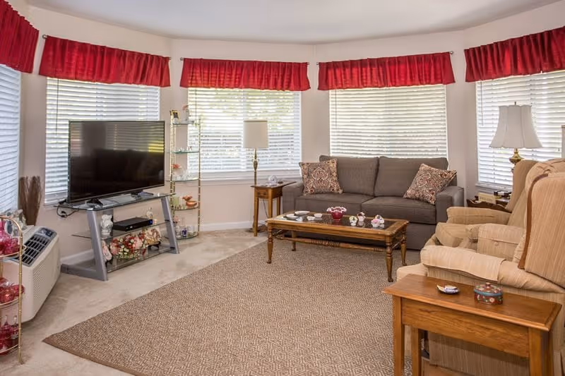 A cozy living room with a large window covered by white blinds and red valances. The room features a gray sofa with patterned pillows, two beige armchairs, a wooden coffee table with decorative items, a TV on a glass stand, and two side tables with lamps. The floor is carpeted with a large area rug.