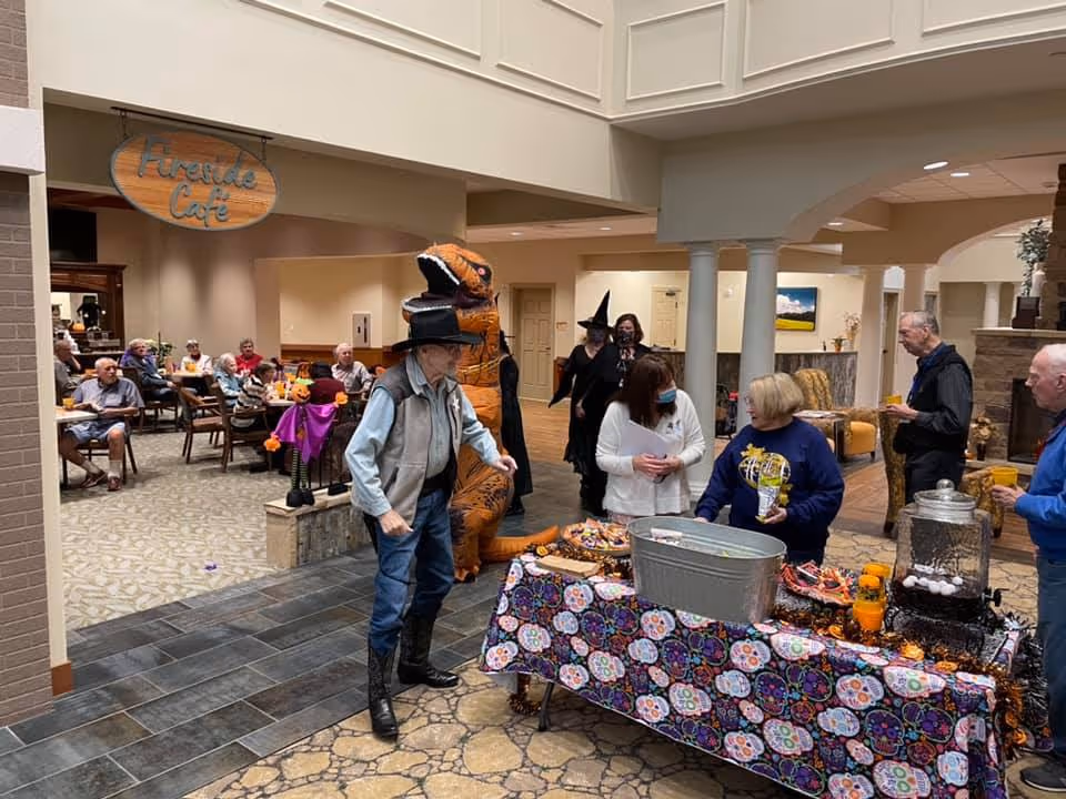 A festive indoor gathering at AHC Knoxville with senior residents and staff. Several people are dressed in Halloween costumes, including a person in a dinosaur costume and another in a witch hat. A table covered with a colorful patterned cloth holds snacks and drinks. In the background, more residents are seated at tables under a sign that reads 'Friends Cafe'.