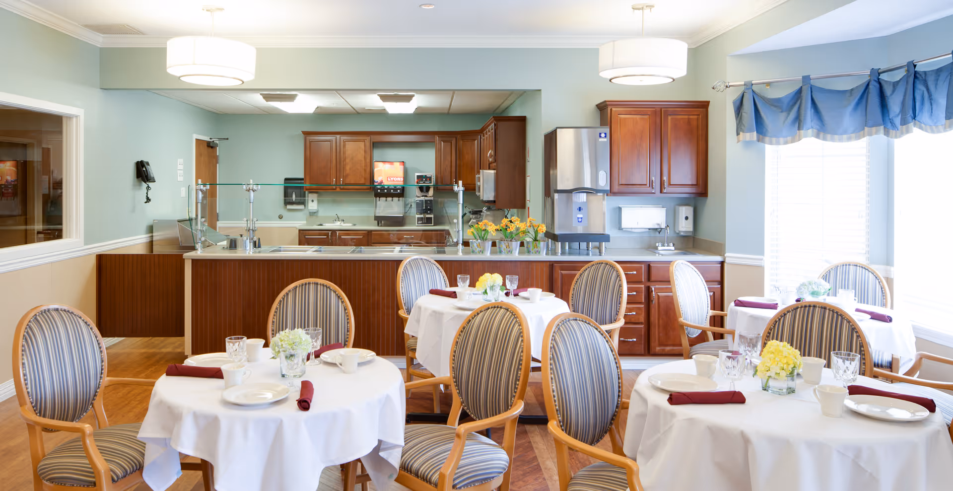 A bright dining room with round tables covered in white tablecloths, each set with plates, cups, and burgundy napkins. The chairs have wooden frames and striped upholstery. In the background, there is a kitchen area with wooden cabinets, a stainless steel water dispenser, and a counter with a glass sneeze guard. The room has light blue walls and large windows with blue valances letting in natural light.