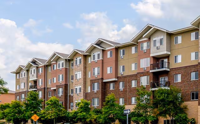 Exterior view of a multi-story senior living facility building with a mix of brick and siding finishes, several windows and balconies, and green trees in front under a partly cloudy sky.
