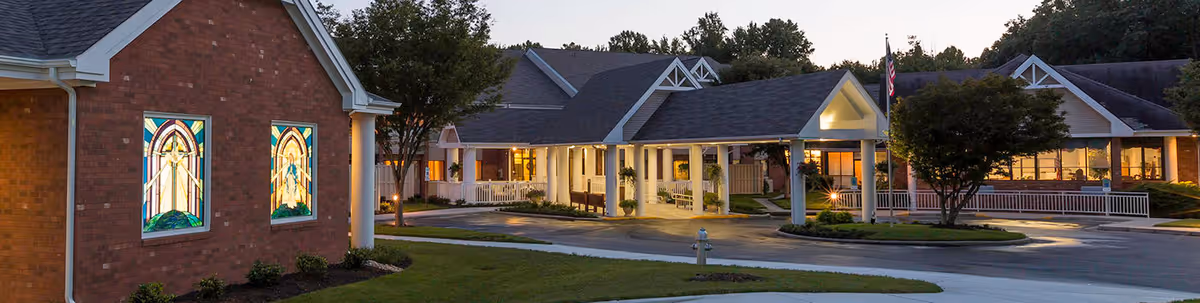 Exterior front of a senior living facility at dusk with a covered entrance, stained-glass windows, and lit interior common areas.