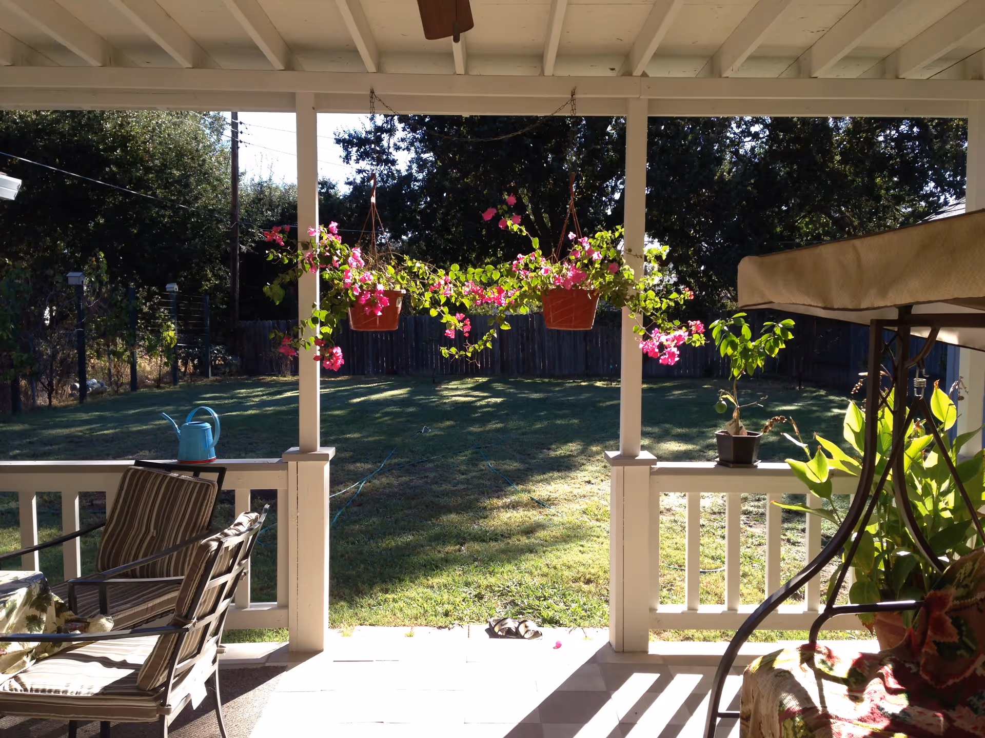 View from a covered porch showing hanging pink flower baskets, patio chairs, potted plants, and a sunny fenced backyard.