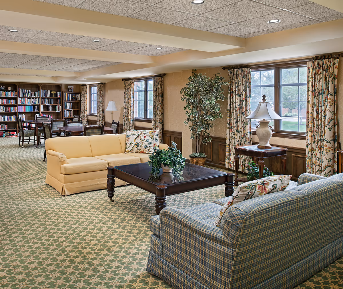 A cozy living room area in a senior living facility with two sofas, a wooden coffee table with a plant, a side table with a lamp, large windows with floral curtains, a potted plant, and bookshelves with tables and chairs in the background.