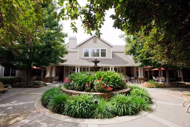 Outdoor courtyard area with a circular garden bed filled with green plants and flowers surrounding a central water fountain. The courtyard is framed by trees and benches, with a two-story building in the background featuring large windows and a covered patio with tables and chairs.