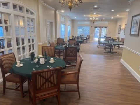 A spacious dining room with multiple round tables covered with green tablecloths, each set with white cups, saucers, and napkins. Wooden chairs with striped cushions surround the tables. The room has large windows allowing natural light, chandeliers hanging from the ceiling, and light-colored walls with white trim.
