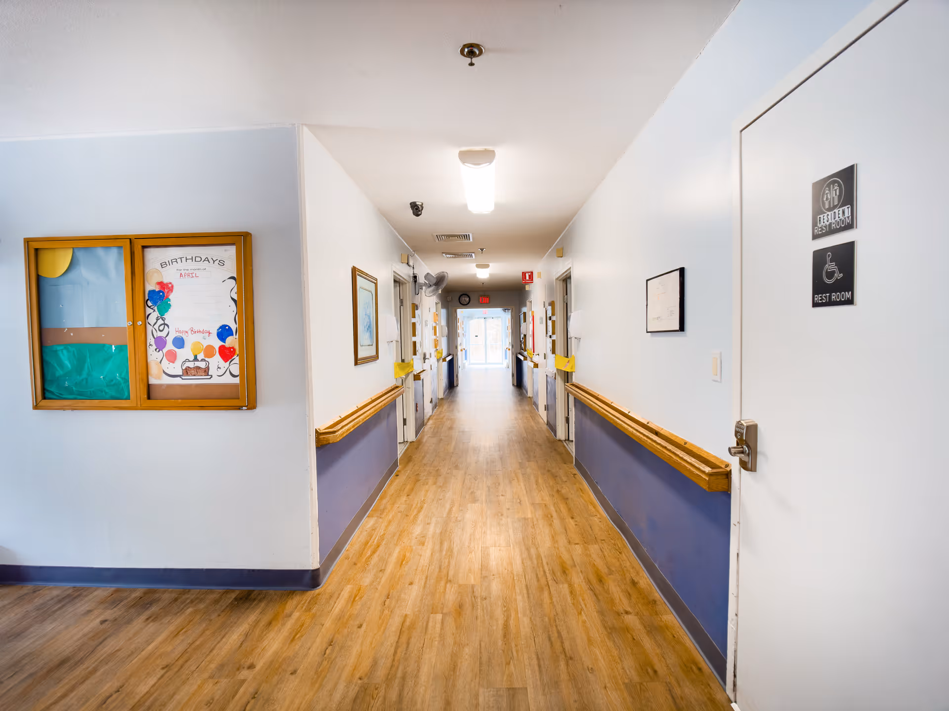 A well-lit hallway in a senior living facility with wooden handrails on both sides, light wood flooring, and white and blue walls. On the right side, there is a door labeled as a resident restroom with accessibility signs. On the left wall, there is a bulletin board with a birthday announcement for April. The hallway extends to a glass door at the far end.