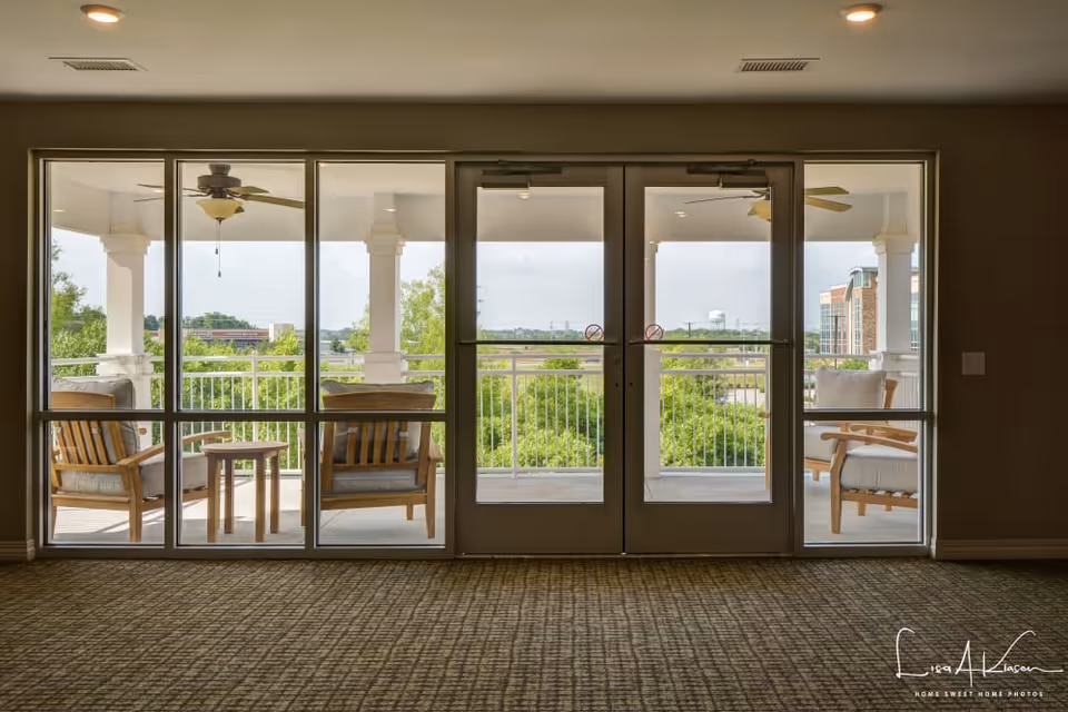 View from inside a room looking out through large glass doors onto a covered balcony with wooden chairs and a small table. The balcony has white railings and columns, ceiling fans, and overlooks greenery and distant buildings.