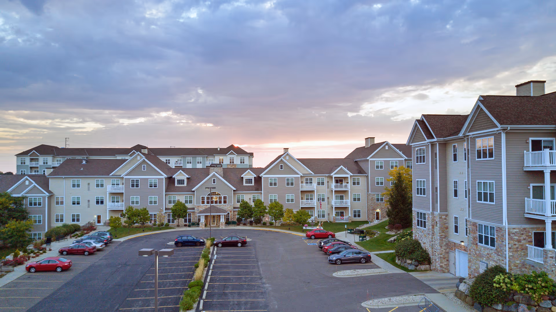 Exterior view of a large senior living facility named All Saints Neighborhood during sunset, showing multiple connected buildings with beige siding and stone accents, balconies, and a parking lot with several cars parked.