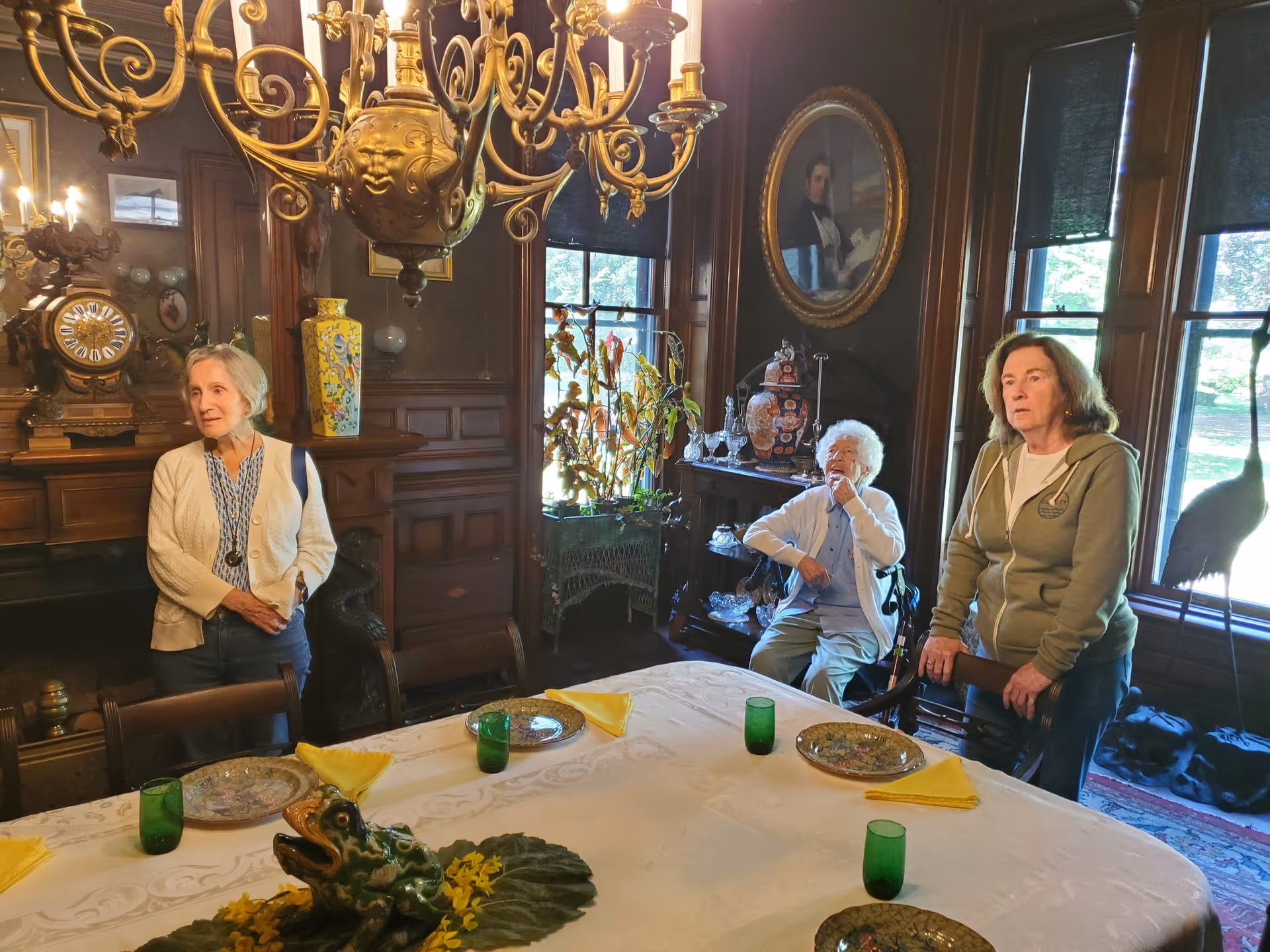 Three elderly women in a richly decorated dining room with dark wood paneling, a large ornate chandelier, and a table set with green glasses, patterned plates, and yellow napkins. The room features antique furniture, a large clock, decorative vases, and a portrait on the wall. Sunlight streams through the windows with dark blinds.