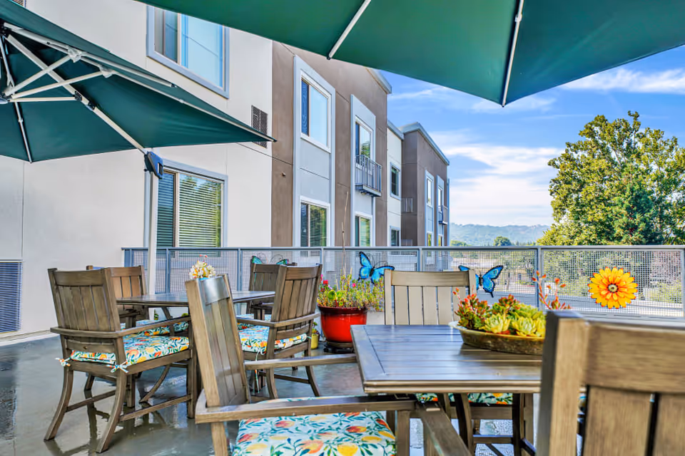 Outdoor patio with wooden tables and chairs under green umbrellas, potted plants and decorative accents on a balcony by a multi-story building.
