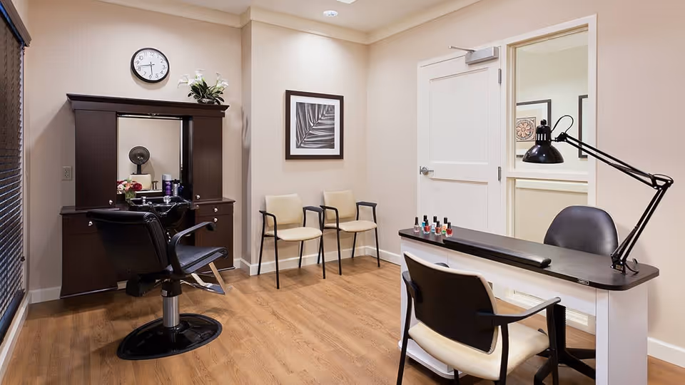 A well-lit salon room with a black salon chair in front of a dark wooden vanity mirror, two beige chairs against the wall, and a manicure table with a black lamp and several bottles of nail polish.