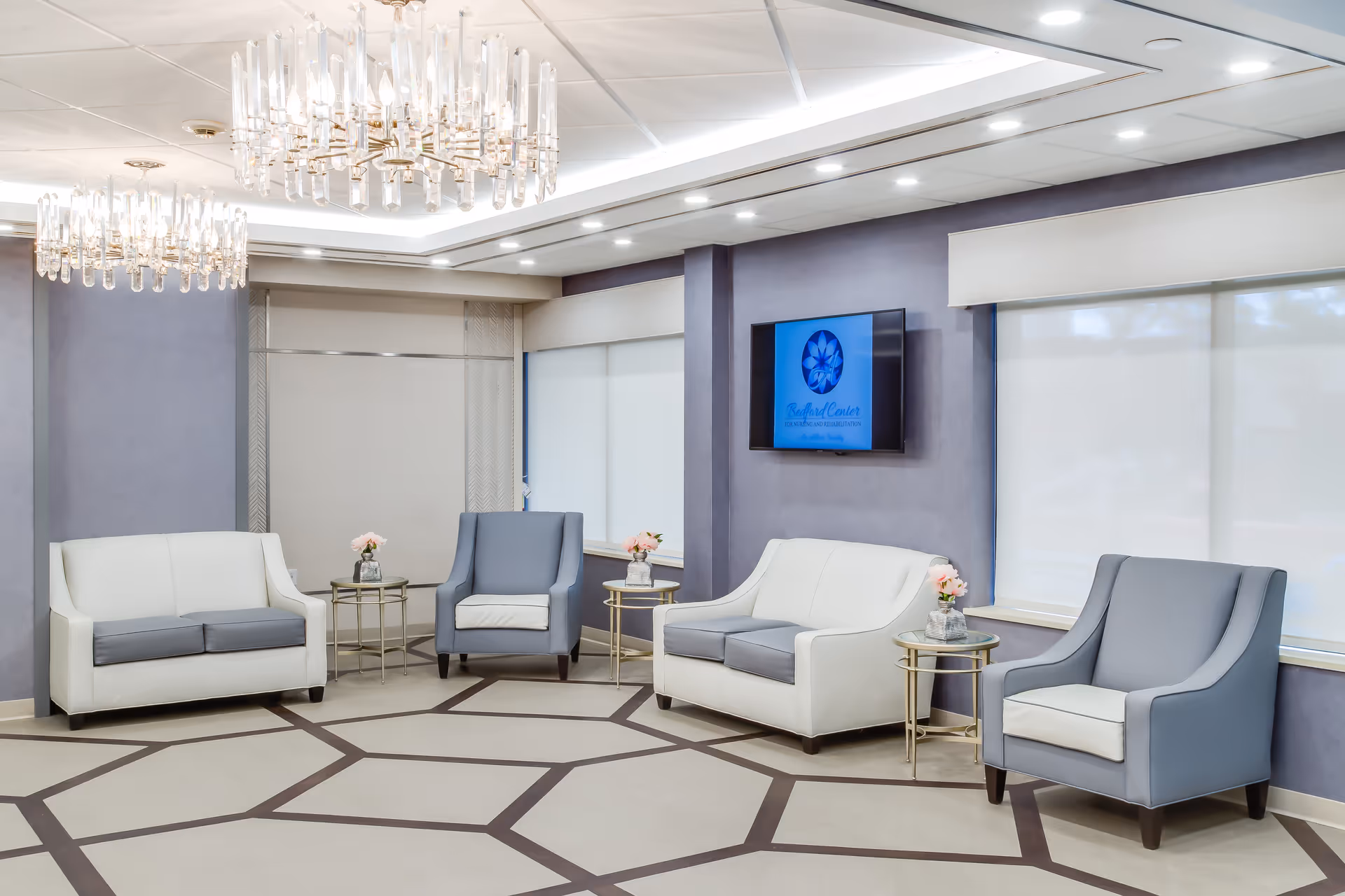 A modern and elegant lounge area with two white and gray sofas, two gray armchairs, small round side tables with flower vases, large windows with white blinds, and two crystal chandeliers hanging from the ceiling. A flat-screen TV mounted on the wall displays the Bedford Center logo.